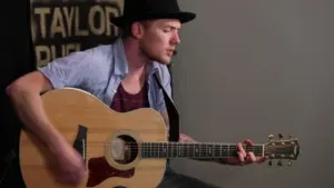 Musician in a black hat playing acoustic guitar, focused on performance, with a backdrop displaying "TAYLOR."