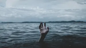 A lone hand reaches out from dark water under a moody sky, evoking a sense of struggle and desperation.