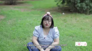 Woman sitting on grass wearing a blue top and headband, speaking directly to the camera in a natural outdoor setting.