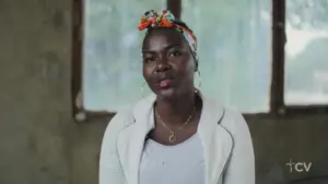 Woman with a colorful headscarf sits indoors, expressing confidence and warmth against a rustic background.