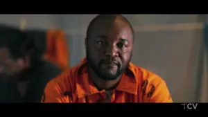 Man in an orange prison jumpsuit sitting indoors, with a serious expression and blurred background of a prison setting.