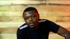 Man in a black shirt with white stripes poses thoughtfully against a rustic wooden background, conveying contemplation.