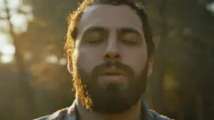 Man with a beard and closed eyes, basking in sunlight amidst a natural backdrop, evoking peace and serenity.