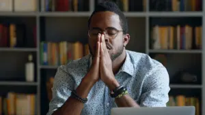 Man in glasses with hands clasped in prayer, focused expression, sitting at a desk with bookshelves in background.