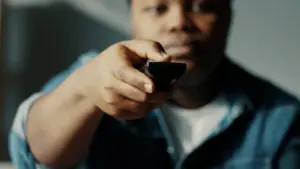 Person pointing a remote control towards the camera, focusing on hands and expression, in a cozy indoor setting.