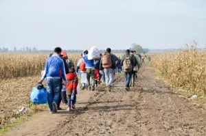 Group of migrants walking through a muddy field, carrying belongings, with blurred landscape in the background.