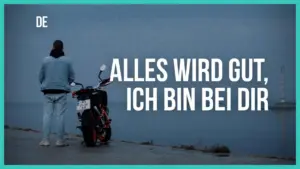 Person standing by a motorcycle near the water, conveying a sense of reassurance with "Alles wird gut, ich bin bei dir."