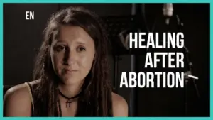 Young woman with dreadlocks sharing her story about healing after abortion, highlighted by a microphone backdrop.