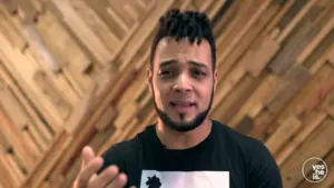 Man with a stylish haircut speaks passionately in front of a wooden backdrop, wearing a graphic t-shirt.