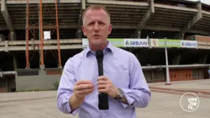 Man holding a microphone outside a stadium, speaking to the camera with construction scaffolding in the background.