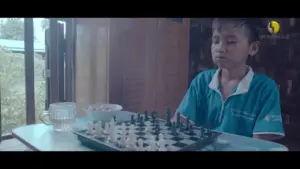 Young boy focused on a chess game, seated at a table with a glass of water and a bowl beside him. Natural light streaming in.