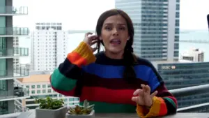 Woman in colorful striped sweater gesturing while sitting at a table with plants, city skyline in the background.