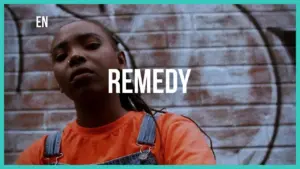 Young woman in orange shirt with denim overalls poses confidently in front of a graffiti wall featuring the word "Remedy."