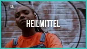 Young woman in orange shirt stands confidently against a graffiti wall, featuring the word "HEILMITTEL" prominently.