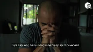 Man with a shaved head sitting indoors, holding hands in prayer, seeking peace and tranquility.