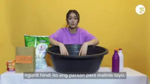 Young woman demonstrating a cleaning process using a large basin, colorful detergent bottles, on a bright yellow backdrop.