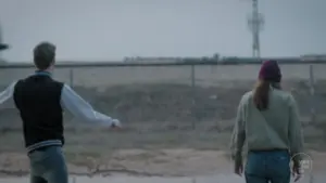 Two people stand in the foreground, looking towards a distant industrial landscape under a cloudy sky.