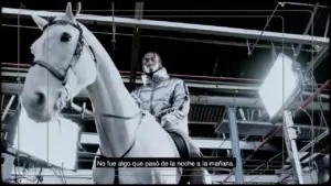 A person in a metallic jacket sits on a horse in a dimly lit industrial setting with bright lights in the background.