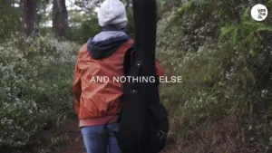 Person walking through a forest path with a guitar case, wearing an orange jacket and gray beanie, text overlay present.