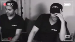 Two men in black shirts sitting in a studio, one laughing and the other covering his face, captured in a candid moment.