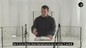 Man speaking passionately while holding an open Bible, surrounded by microphones and a minimalist white backdrop.