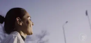 Smiling athlete with a bun, enjoying a sunny outdoor moment against a clear sky and blurred streetlights in the background.