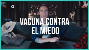 A surprised man sitting on a couch, holding a remote, with bold text reading "Vacuna contra el miedo" in a dark room.