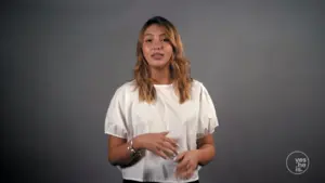 Smiling woman in a white top stands against a gray background, gesturing while delivering a message or presentation.