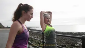 Two women exercising outdoors, one stretching while the other walks along a scenic coastal path. Bright athletic wear.