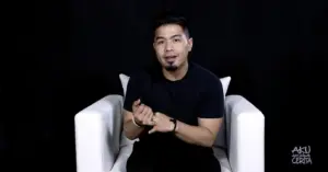 Person in a black shirt sitting on a white chair against a black background, gesturing with hands while speaking.