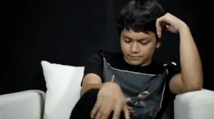 Young man in a black T-shirt resting his chin on his hand, seated on a couch against a dark backdrop.