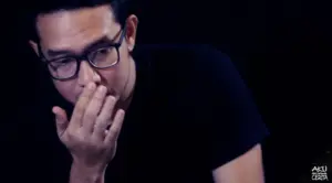 Man in glasses contemplatively touching his face against a dark background, expressing deep thought or concern.