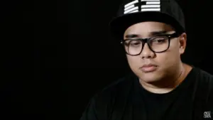 Young man wearing a black cap and glasses, with a thoughtful expression against a dark background.