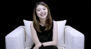 Young woman seated on a white couch, smiling against a black backdrop, exuding warmth and approachability.