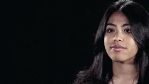 Young woman with long black hair, looking thoughtfully to the side against a dark background.