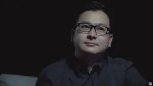 A contemplative man in glasses looks thoughtfully into the distance against a dark background.