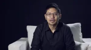 Man wearing glasses sitting on a white couch in a dark room, smiling and looking directly at the camera.