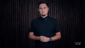 Man in a black shirt stands against a dark wooden backdrop, conveying a confident and engaging attitude.