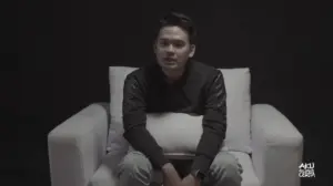 Young man sitting on a white couch against a dark background, looking directly at the camera with a contemplative expression.