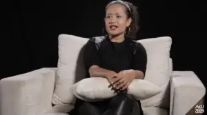 Woman sitting on a couch with a pillow, smiling and engaging in conversation against a dark background.