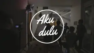 Logo "Aku Dulu" in a creative workspace with people collaborating and modern decor in the background.