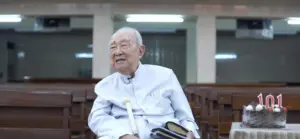 Elderly man celebrating his 101st birthday with a cake and books in a simple indoor setting, smiling joyfully.