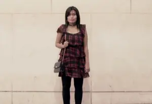 Young woman in a plaid dress and black leggings stands against a beige wall, holding a small purse.