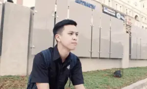 Young man in a black shirt sitting outdoors, looking thoughtfully into the distance against a city backdrop.