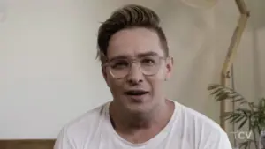 Person with glasses and styled hair, wearing a white t-shirt, seated in a casual indoor setting.