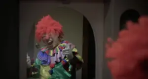 A cheerful clown in vibrant costume and orange wig smiles and points playfully in a lively indoor setting.