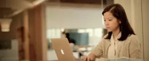 Young woman focused on laptop in modern office setting, showcasing productivity and a collaborative work environment.