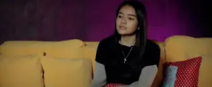 Teen girl sitting on a yellow couch, wearing a black shirt and silver necklace, with a colorful background.