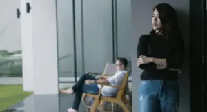 A woman in a black top stands thoughtfully, while a man relaxes in a chair, showcasing modern interior design and introspection.