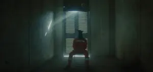 Person in a red outfit sitting in a dimly lit room, facing a door with dramatic light streaming through slatted blinds.